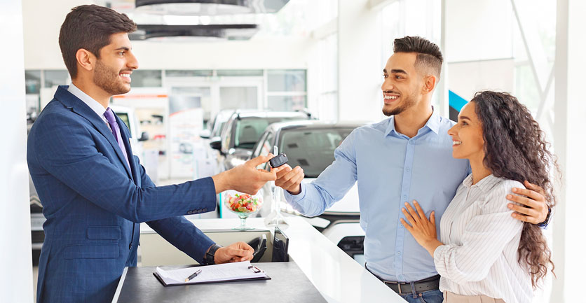 Pareja de jóvenes felices recibiendo las llaves de su nuevo automóvil entregadas por un joven empresario